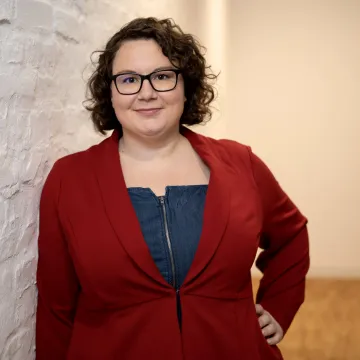 Maria Hinz, Frau mit braunen Locken, Brille und rotem Blazer, die an einer weißen Wand lehnt